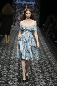 Laufsteg-Model trägt knielanges, wallendes Kleid mit blauem Floralmuster im Stil der 60er.